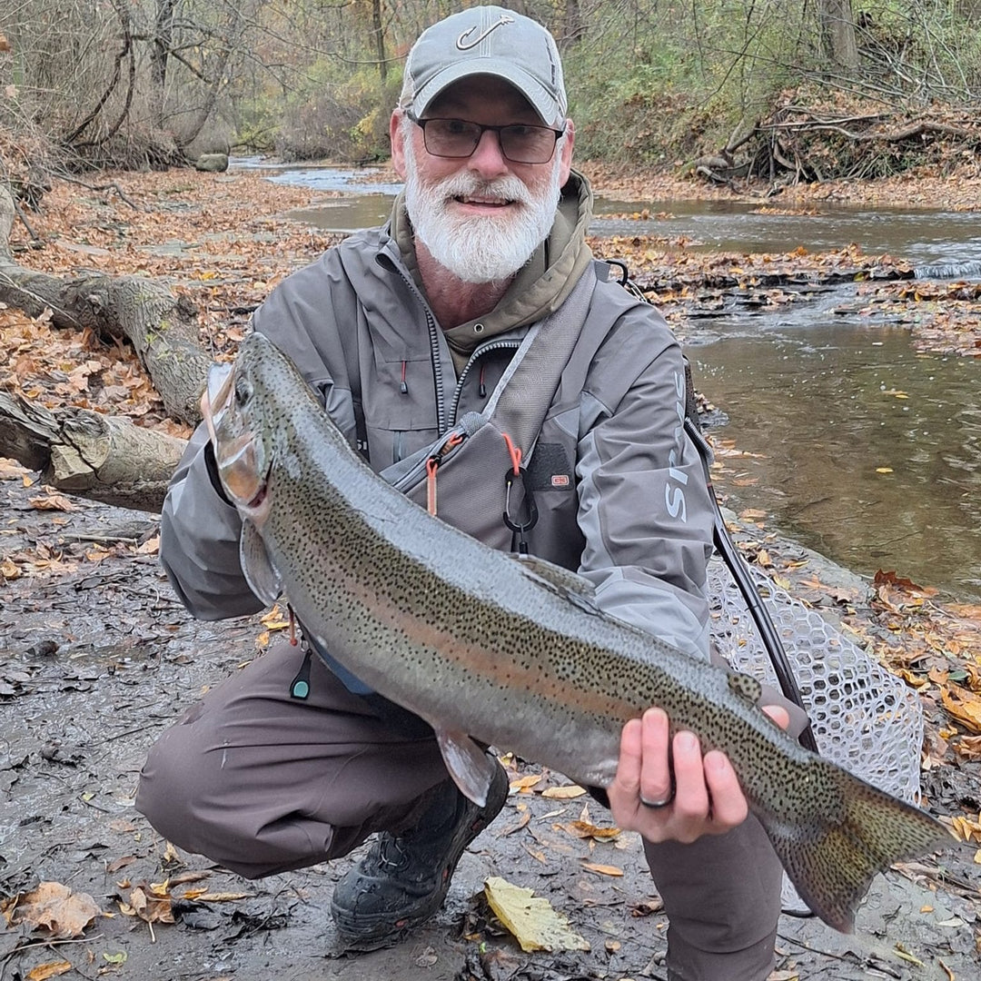 An angler holding a large steelhead trout he caught with a Wasatch Tenkara Rods - RodZilla, with a big smile, standing on a muddy riverbank surrounded by autumn foliage and a peaceful stream in the background.