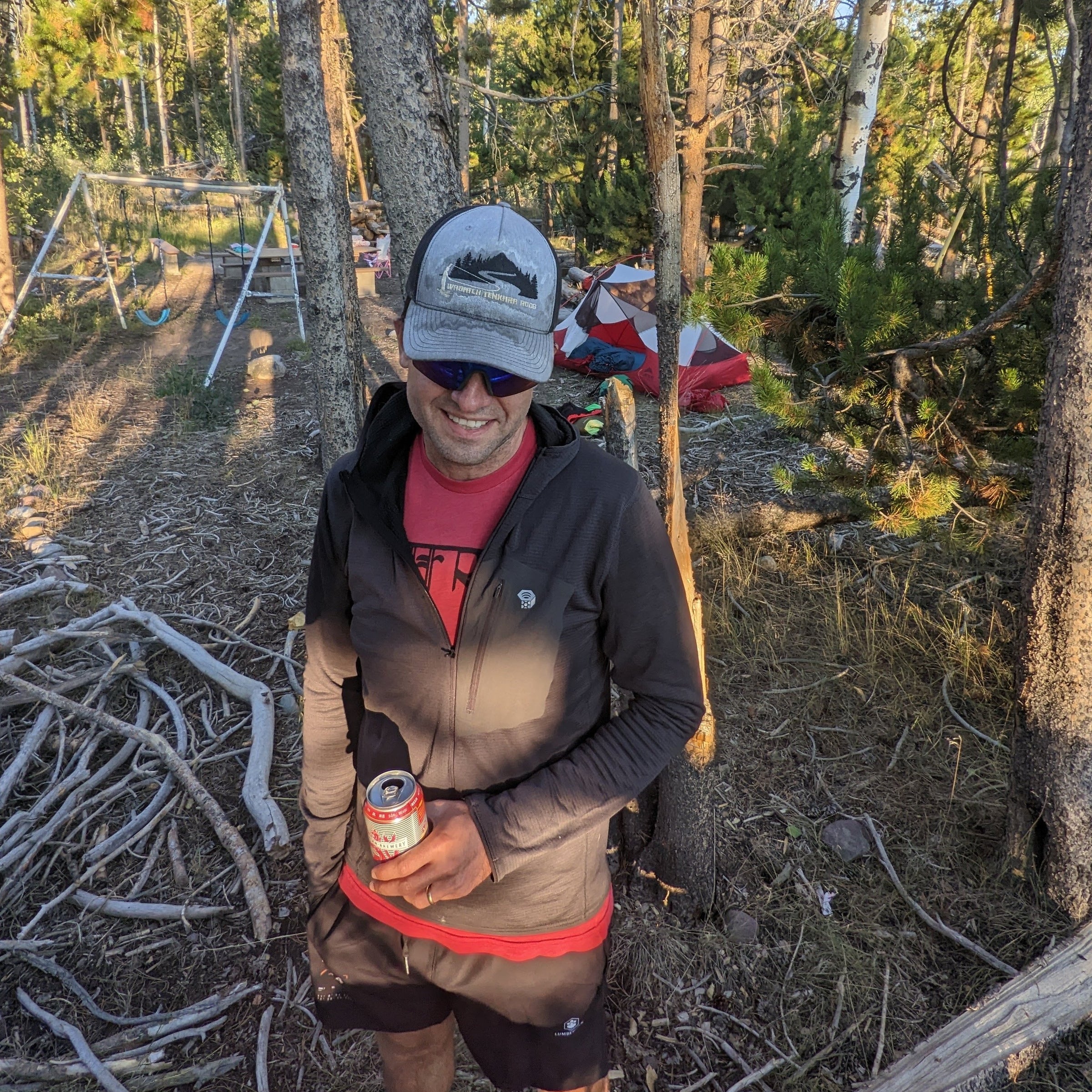 A man stands in a wooded campsite wearing a Wasatch Tenkara Rods cap with a design of a man fishing, sunglasses, and outdoor gear while holding a beverage can. In the background, a hammock, a tent, and scattered camping gear are visible among the trees, creating a relaxed and adventurous atmosphere in the wilderness.