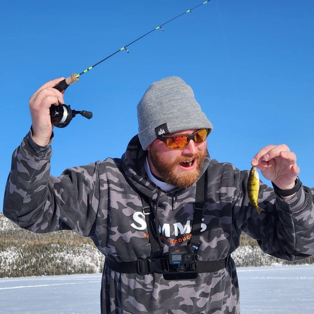 A bearded angler in a camouflage hoodie and gray beanie holding up a small yellow perch and a Wasatch Tenkara Rods - Ice Breaker ice fishing rod with a cork handle, smiling enthusiastically against a bright blue sky on a frozen lake.