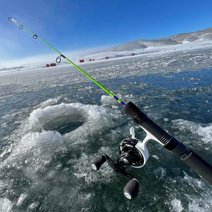 A bright green Wasatch Tenkara Rods - The Joker ice fishing rod with a spinning reel set up for ice fishing near a freshly drilled hole in a frozen lake, surrounded by a snowy landscape and distant red ice fishing shelters under a clear blue sky.