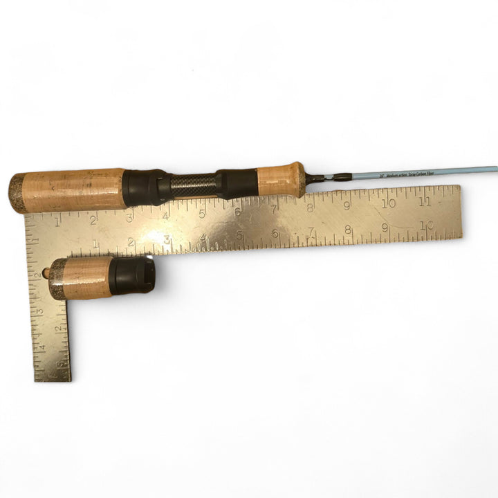 A Wasatch Tenkara Rods - Winterhawk ice fishing rod handle made of cork placed alongside a metal ruler for size comparison, with a sleek blue rod section visible, displayed on a white background.