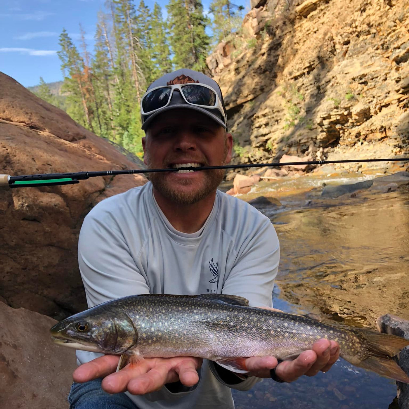 A smiling angler holds a vibrant brook trout with both hands while a Wasatch Tenkara Rods - Darth Quattro is held between his teeth. The setting features a clear, rocky stream surrounded by tall cliffs and evergreen trees under a sunny blue sky, capturing a peaceful moment in nature.