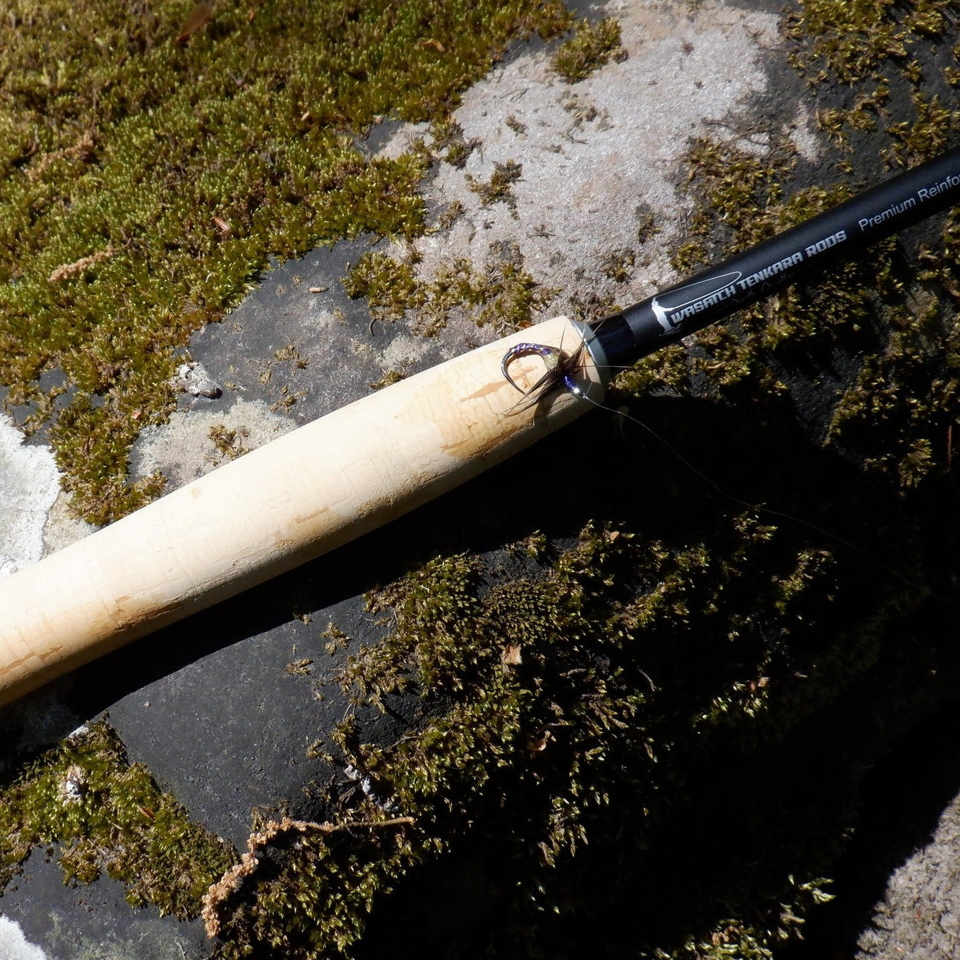 A close-up of a cork-handled Wasatch Tenkara Rods - Darth Quattro fishing rod with a small Kebari fishing fly attached, resting on a moss-covered rock. The bright sunlight highlights the texture of the rod and the natural surroundings, creating a serene outdoor fishing scene.